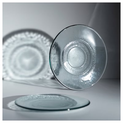 Three transparent glass plates in various positions on a gray surface, showcasing their unique, textured design and recycled glass composition.