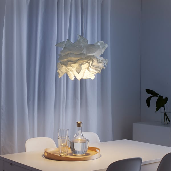 KRUSNING lamp above dining table with glasses and vase, soft white light, surrounded by chairs, window framed by curtains.