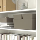 A white bookshelf displays KUGGIS storage boxes and books. The boxes organize small items, with one stackable lid-optional.