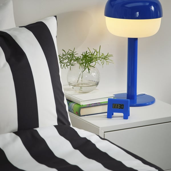 A bedroom nightstand with a blue table lamp, clock, books, and a striped pillow. The digital clock, KUPONG, has an increasing alarm function.