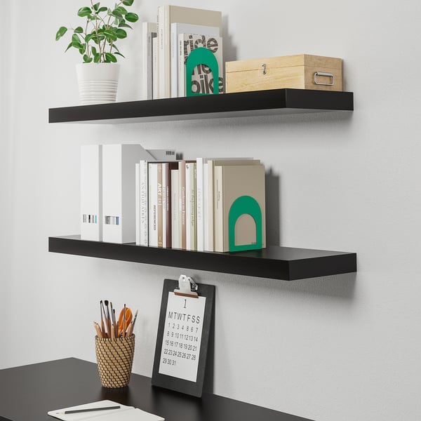 BLACK wall-mounted LACK shelves holding books, plant, and decor; desk below with calendar and pens.