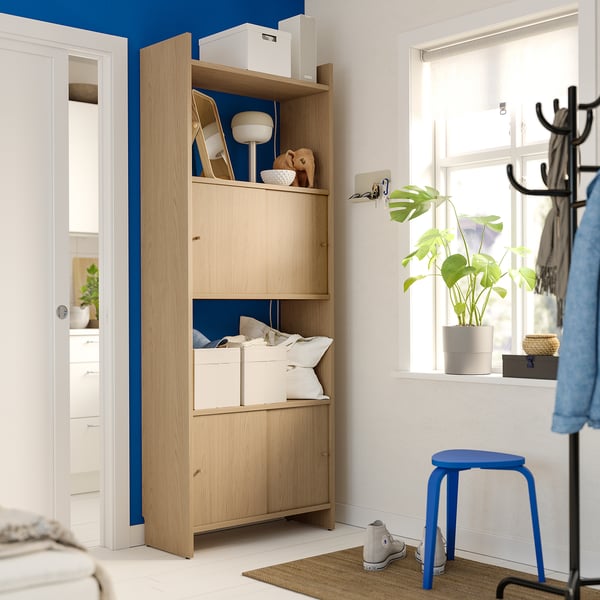 An LÅDMAKARE storage unit in light wood against a blue wall. It has open shelves and closed cabinets with a minimalist design.