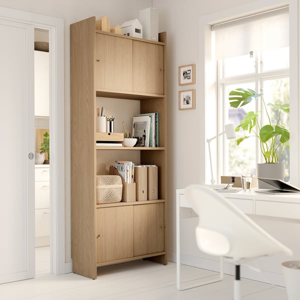 Modern light wood bookcase with open shelves and closed cabinets. Held against wall, bright room with white furniture.