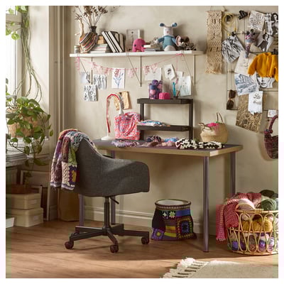 Craft room with LAGKAPTEN desk, shelves with yarn, knitted items, and decorative objects. Grey chair, plant, and wall decorations.