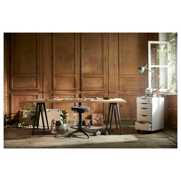 Modern desk setup with wooden top, black legs, stool, lamp, and storage against wooden walls with a window.