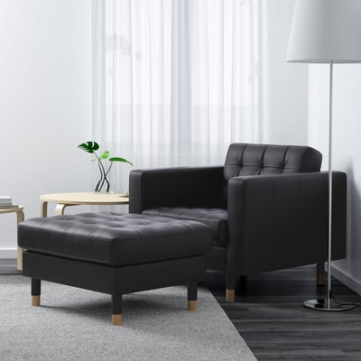 Elegant living room with black LANDSKRONA chair & ottoman, sleek lines, beside tall lamp, light wood side table with plant.