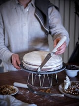 A person cuts a white frosted cake with a white-handled knife on a metal stand. Plates of nuts and cookies surround the cake.