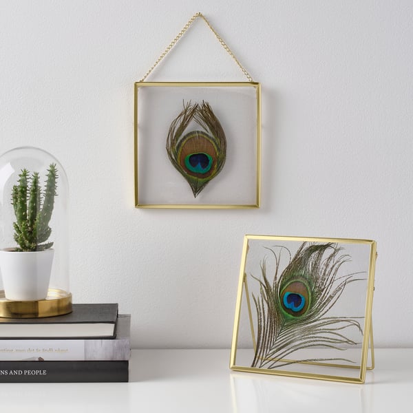 Two gold-framed peacock feather artworks displayed; one hangs on the wall, the other stands on a surface, showcasing interchangeable motifs.