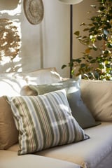 Bed with green and white striped pillow, beige pillows, woven decor on wall, and tall plant.