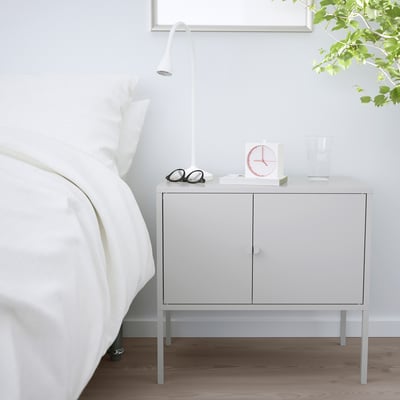 A white IKEA LIXHULT storage cabinet next to a bed. Its compact, with two doors, and supports a lamp, clock, glasses, and a vase.