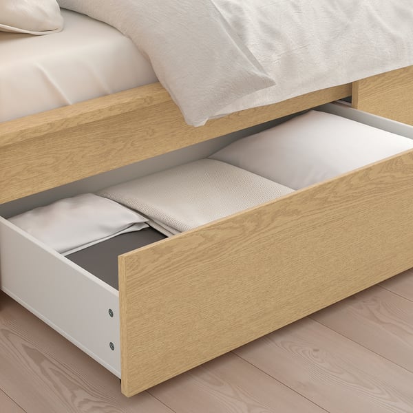A light wooden bed with two large storage drawers open, revealing blankets inside. The drawers have casters for smooth rolling.