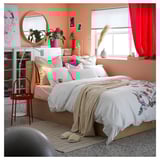 Peach-walled bedroom with white bed, gold blanket, colorful pillows, red stool, shelves, plants, large window with red curtains.