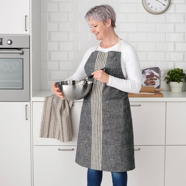 A woman wearing a gray MARIATHERES apron is whisking in a bowl in a kitchen. The apron has pockets and a loop for hanging tools.