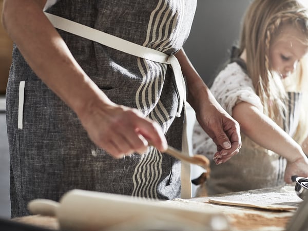 Two people wearing MARIATHERES aprons prepare food. Aprons are linen/cotton blend with pockets and loops. One person is cutting dough.