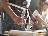 Two people wearing MARIATHERES aprons prepare food. Aprons are linen/cotton blend with pockets and loops. One person is cutting dough.
