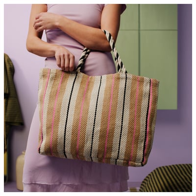A person holding a woven bag, featuring beige, black, and pink diagonal stripes, with braided handles.