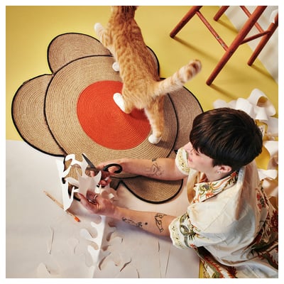 Person sitting, cutting paper with scissors; cat jumping on braided jute rug with red center by MÄVINN.