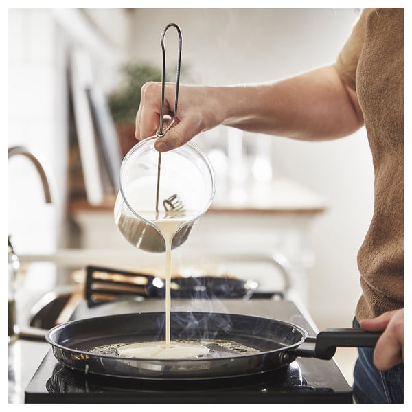Person pouring batter into a black skillet on a stovetop. The pan is in a kitchen setting with a window in the background.