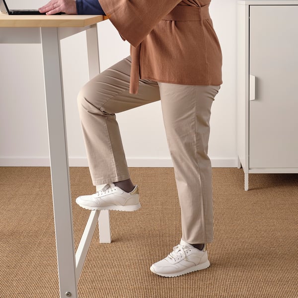 Person standing at tall table with footrest, wearing casual attire and white sneakers.