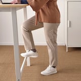 Person standing at tall table with footrest, wearing casual attire and white sneakers.