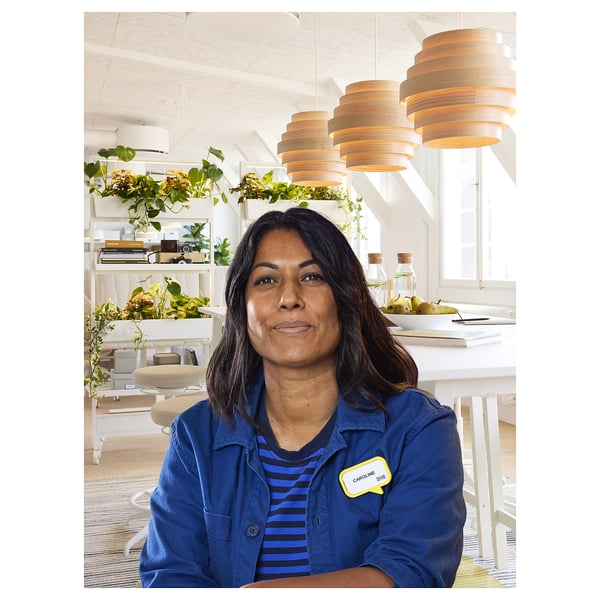 A woman in blue jacket sits at a white desk with three pendant lights and green plants in the background.