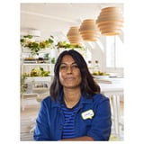A woman in blue jacket sits at a white desk with three pendant lights and green plants in the background.