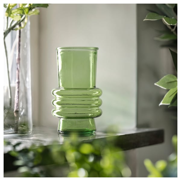 Green, ribbed glass vase on a reflective surface, surrounded by plants.