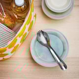 A stainless steel spork, combining spoon and fork in one, sits on a light blue plate. Ideal for travel and outdoor activities.
