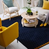 Bright living room with blue NÖVLING rug, yellow chair, wooden coffee table, and wicker chair.