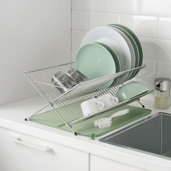 Metal dish drainer on counter with dishes, cups, and cutlery, over green mat, next to sink.