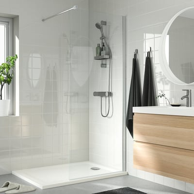 Modern shower with white tiles, glass doors & anti-slip tray. Safety glass, wooden vanity, plant & slippers.