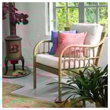 Cozy indoor sitting area with wicker chair, colorful pillows, plants, and a stove heater on a wooden floor.