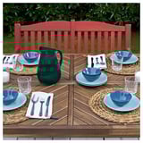 Outdoor table set with blue plates, bowls, and glassware, featuring a red bench with vertical slats and curved backrest, perfect for garden dining.