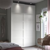 Modern bedroom with white PAX wardrobe, hanging plants, wicker chair, bed with striped blanket, light gray walls, and city view window.