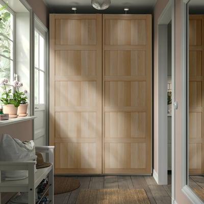 Modern, light wood PAX wardrobe with sliding doors stands tall, filling hallway corner. Sized larger than nearby bench.