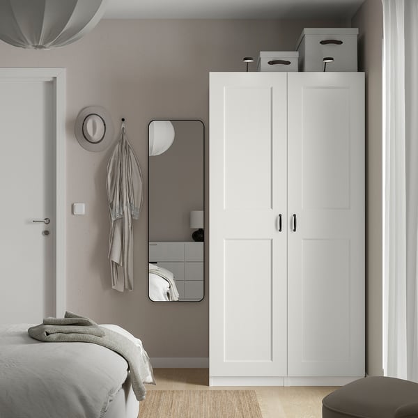 Modern bedroom with white PAX wardrobe, bed, mirror, and gray hat on hooks. Minimalist design, clean lines.