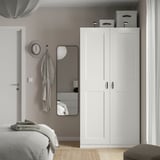 Modern bedroom with white PAX wardrobe, bed, mirror, and gray hat on hooks. Minimalist design, clean lines.