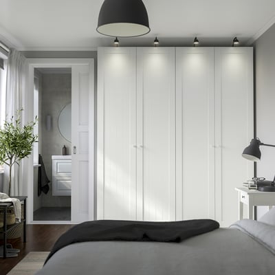Modern bedroom with large white PAX closet, gray bed, plant, and desk lamp. Soft lighting highlights the wardrobes tall, sleek design.