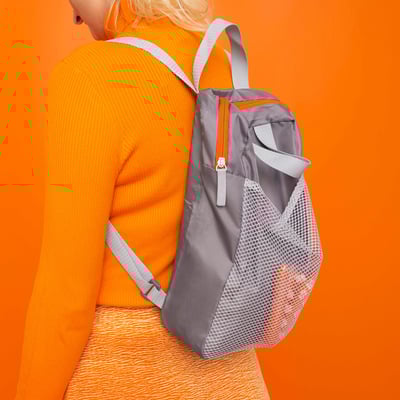 Person wearing orange knitwear, carrying grey PIVRING backpack with mesh pocket against bright orange background.