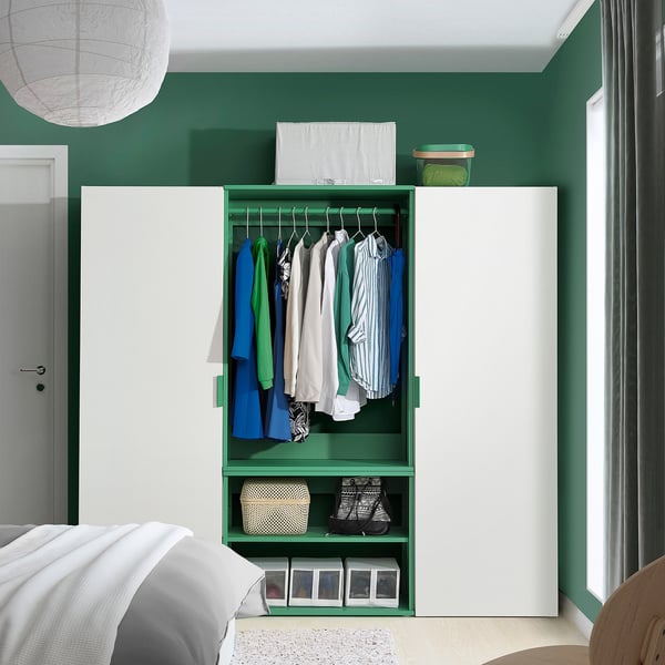 A green PLATSA storage unit holds clothes and boxes in a bedroom next to a bed, fitting beneath a ceiling light.
