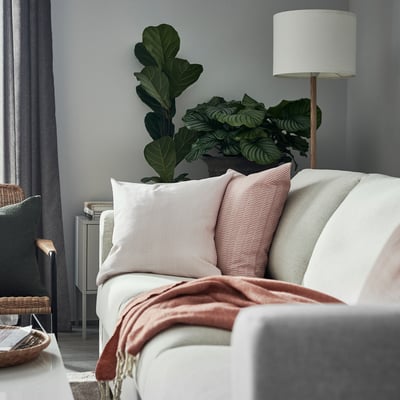 Cozy living room with white couch, pink throw pillows, red blanket, lush green plants, tall lamp, and woven basket.