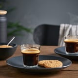 Two small glasses of steaming espresso on plates with cookies beside them.