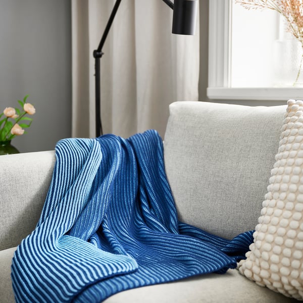 Blue PRAKTFJÄRIL blanket on white sofa, larger than the white pompom-filled pillow.
