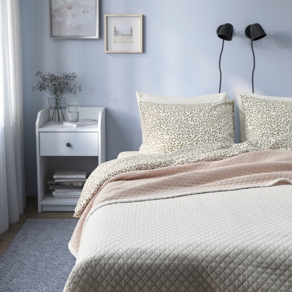 White quilted bedspread with pillows, nightstand with books and vase.