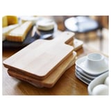 Stacked wooden cutting boards on a table, one with a handle, and a small white bowl.