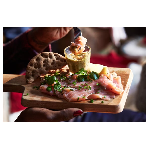 A wooden cutting board displays smoked salmon, bread, and a small cup of sauce. A hand holds a piece of salmon dipped in sauce.