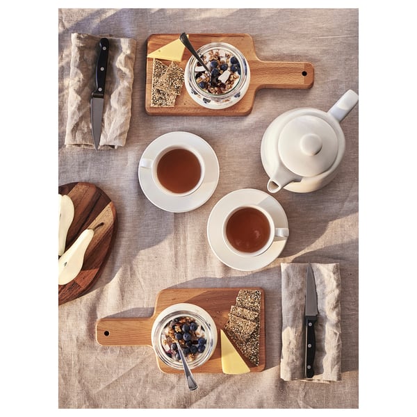 A wooden cutting board and accessories for food prep. Includes a white knife and spoons. Displayed are two saucers with tea.