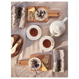 A wooden cutting board and accessories for food prep. Includes a white knife and spoons. Displayed are two saucers with tea.