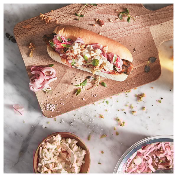 Wooden cutting board with a sandwich topped with onions and sauce, placed on a marble surface with bowls of onions and sauce.