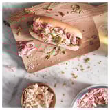 Wooden cutting board with a sandwich topped with onions and sauce, placed on a marble surface with bowls of onions and sauce.